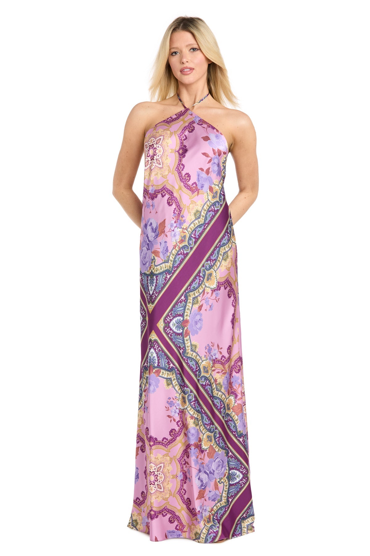 A woman wearing the Paisley Floral Printed Maxi Dress: Morgan 19309, featuring a sleeveless halter neckline and a vibrant pink and purple floral pattern, stands against a plain white background.